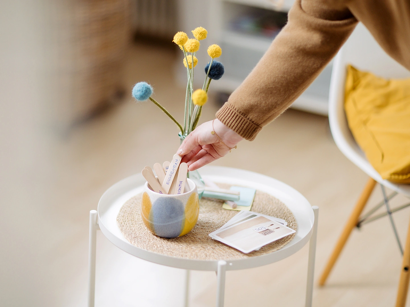 Situations Foto von einem Beistelltisch auf dem eine Tasse mit Holzstäbchen und verschiedenen Worten darauf steht. Eine Hand greift nach dem Holzstäbchen mit dem Wort "Essregeln"
