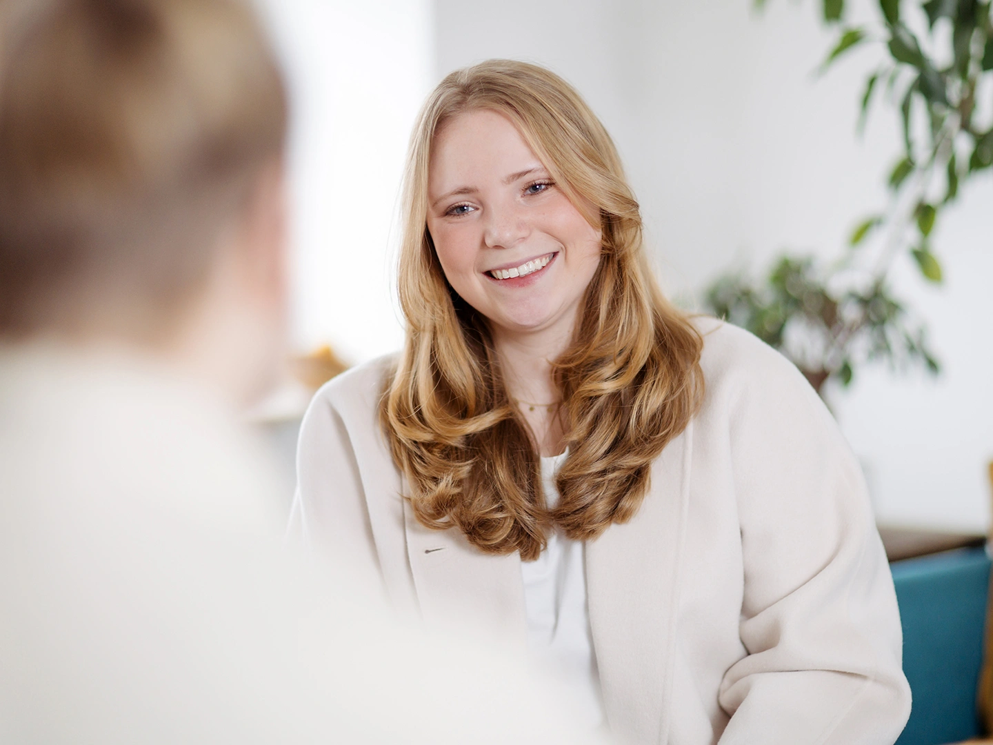 Halbkörper Portrait von Lara Barfuß lächelnd in einer Therapie Situation. Unscharf im Vordergrund links ist die Schulter eine:r Klient:in erkennbar.